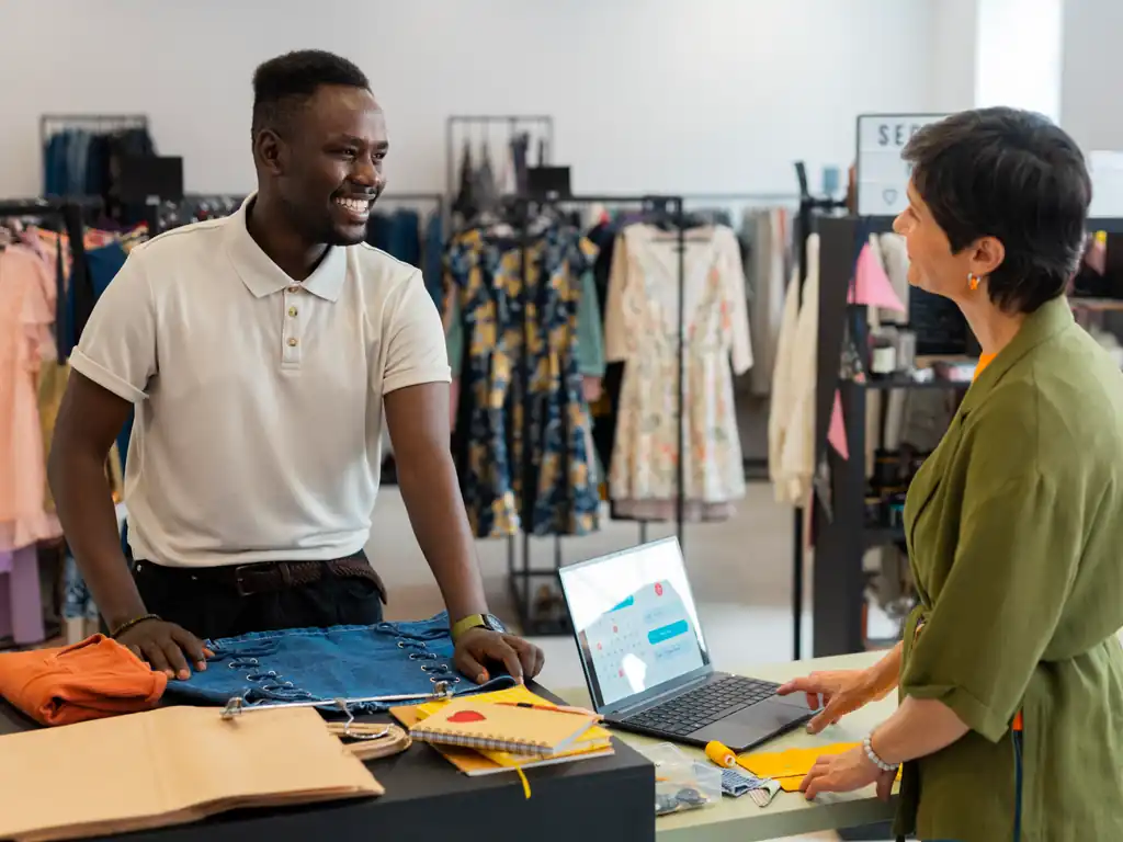A retail shop assistant serving a customer at the counter with a laptop point-of-sale in a clothing store.
